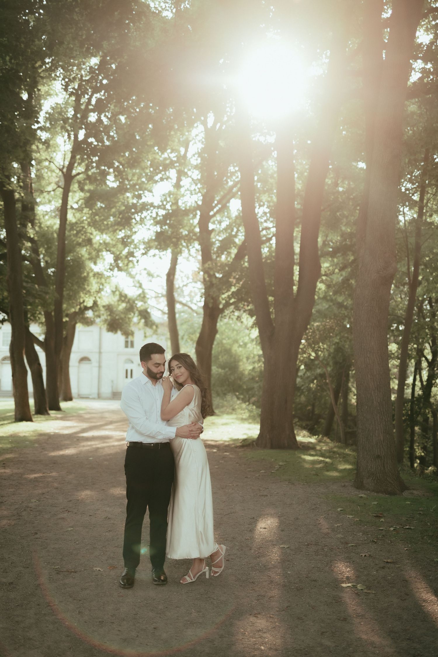 A couple embraces on a sunlit forest path with sunbeams filtering through tall trees on a summer afternoon.