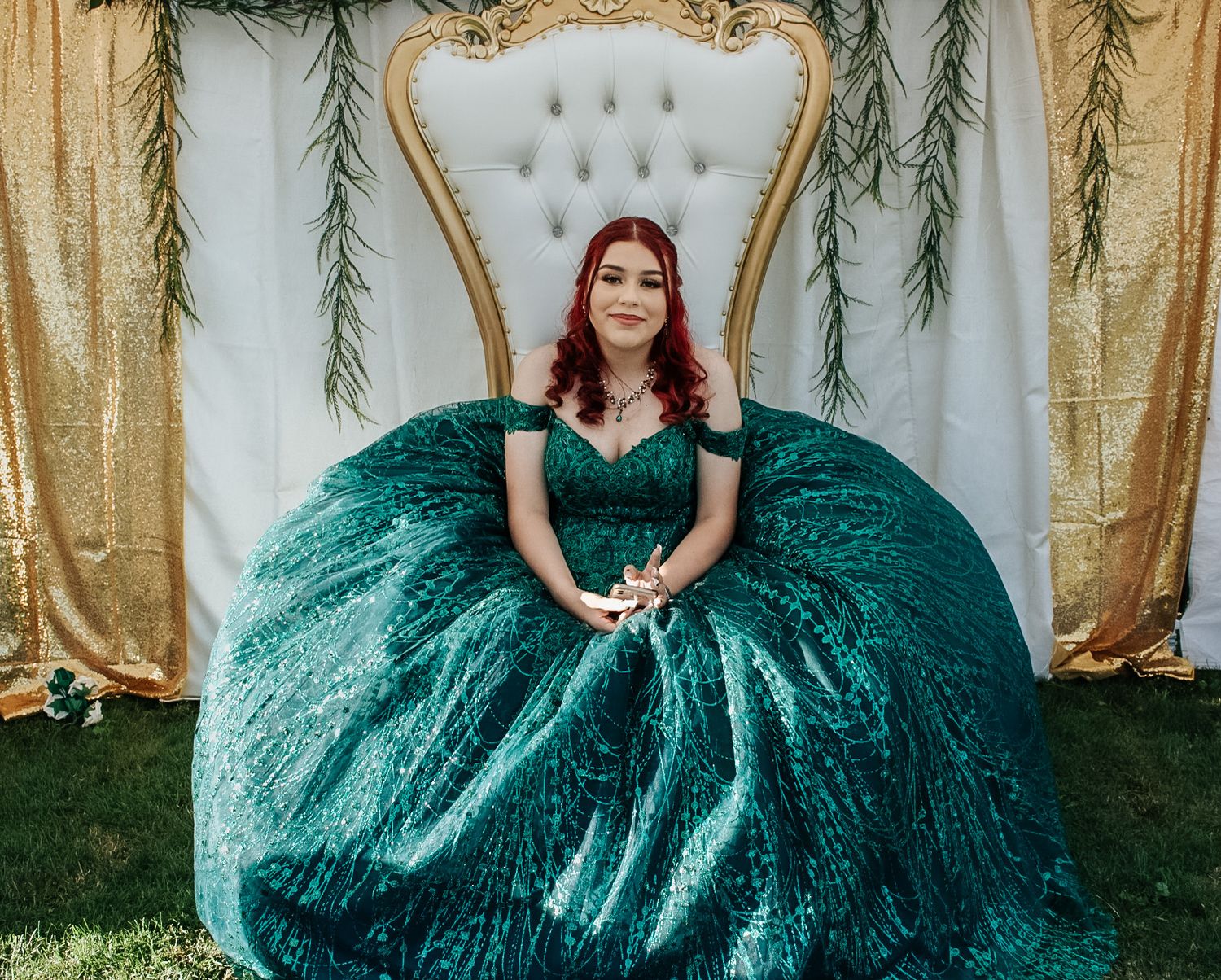Woman in stunning emerald green sequin ball gown sitting on ornate white and gold throne chair for quinceañera.