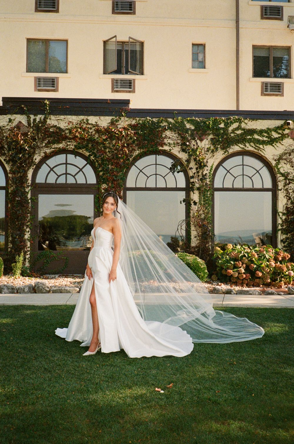 A flowing white wedding dress with a long veil photographed in a garden setting with arched windows.