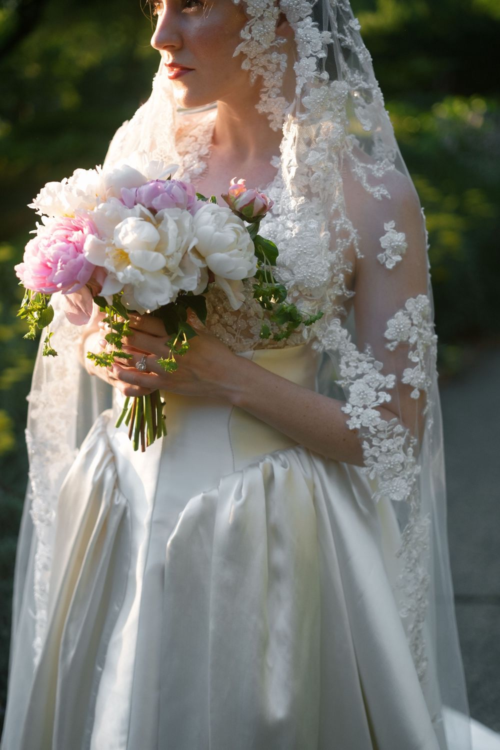 Bride in lace veil and white gown holding pink and white peony bouquet outdoors.