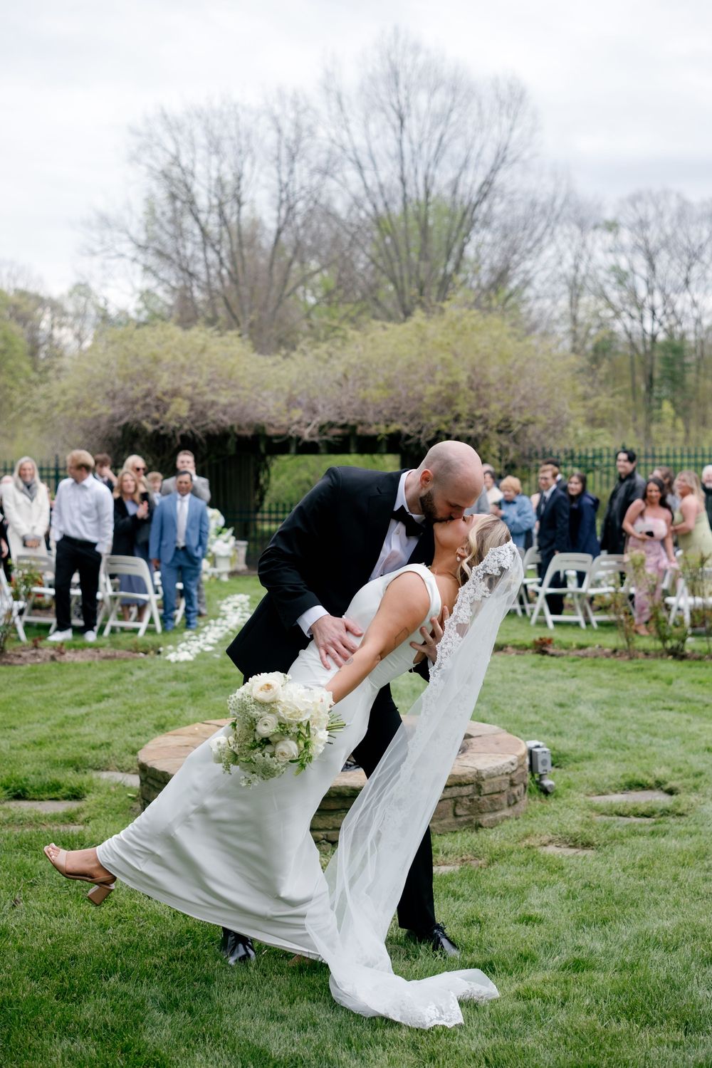 Groom dips bride for a romantic kiss after outdoor wedding ceremony on lush green lawn with guests applauding.