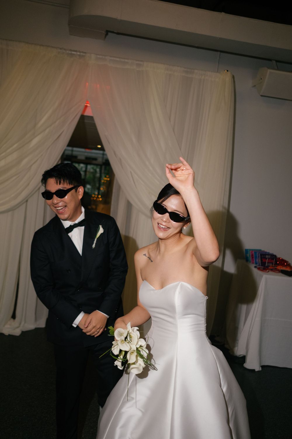 Smiling bride in white strapless gown with sunglasses holds bouquet beside groom in black tuxedo at wedding reception.