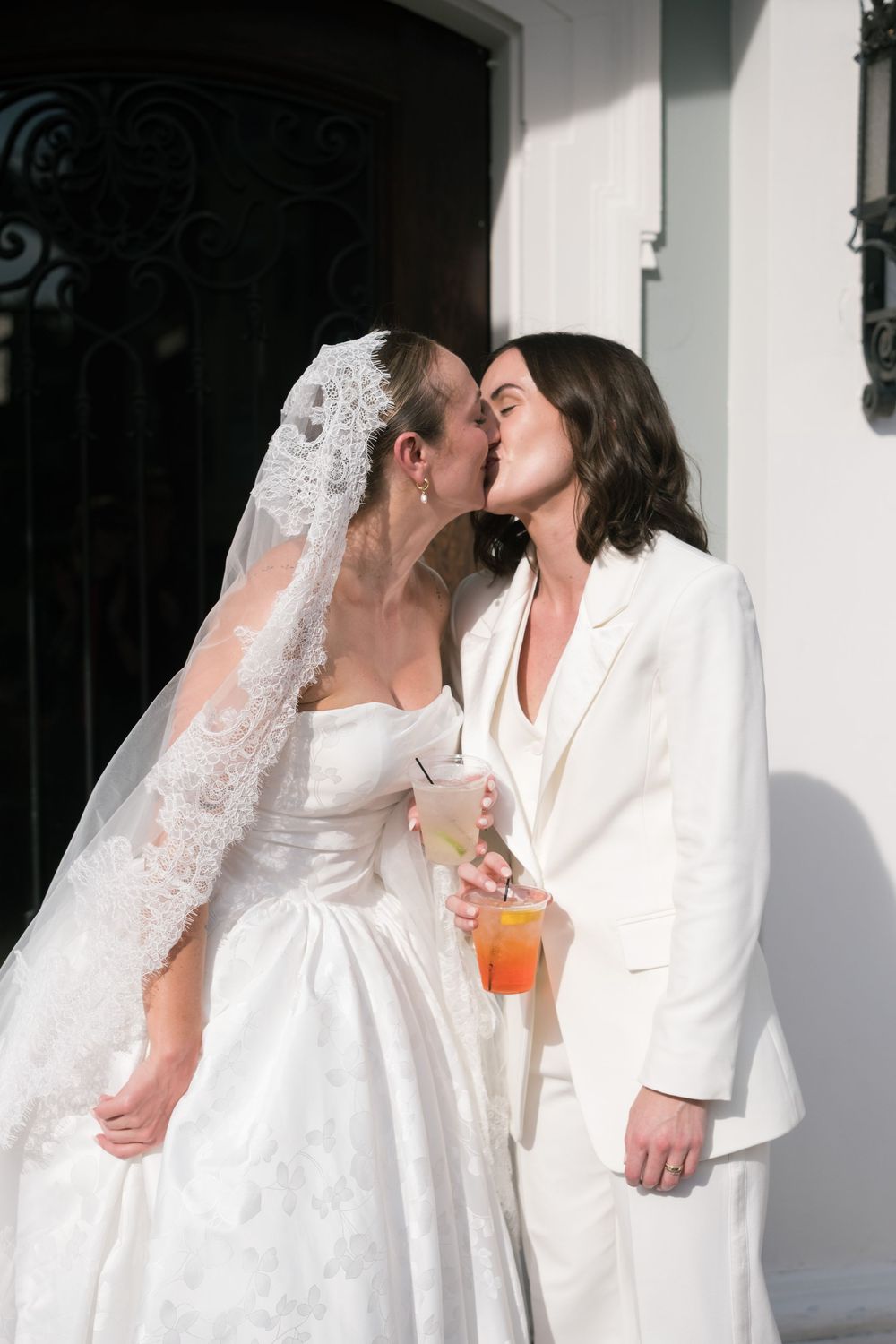 Two people in white wedding attire share a kiss while holding cocktails against a white wall.