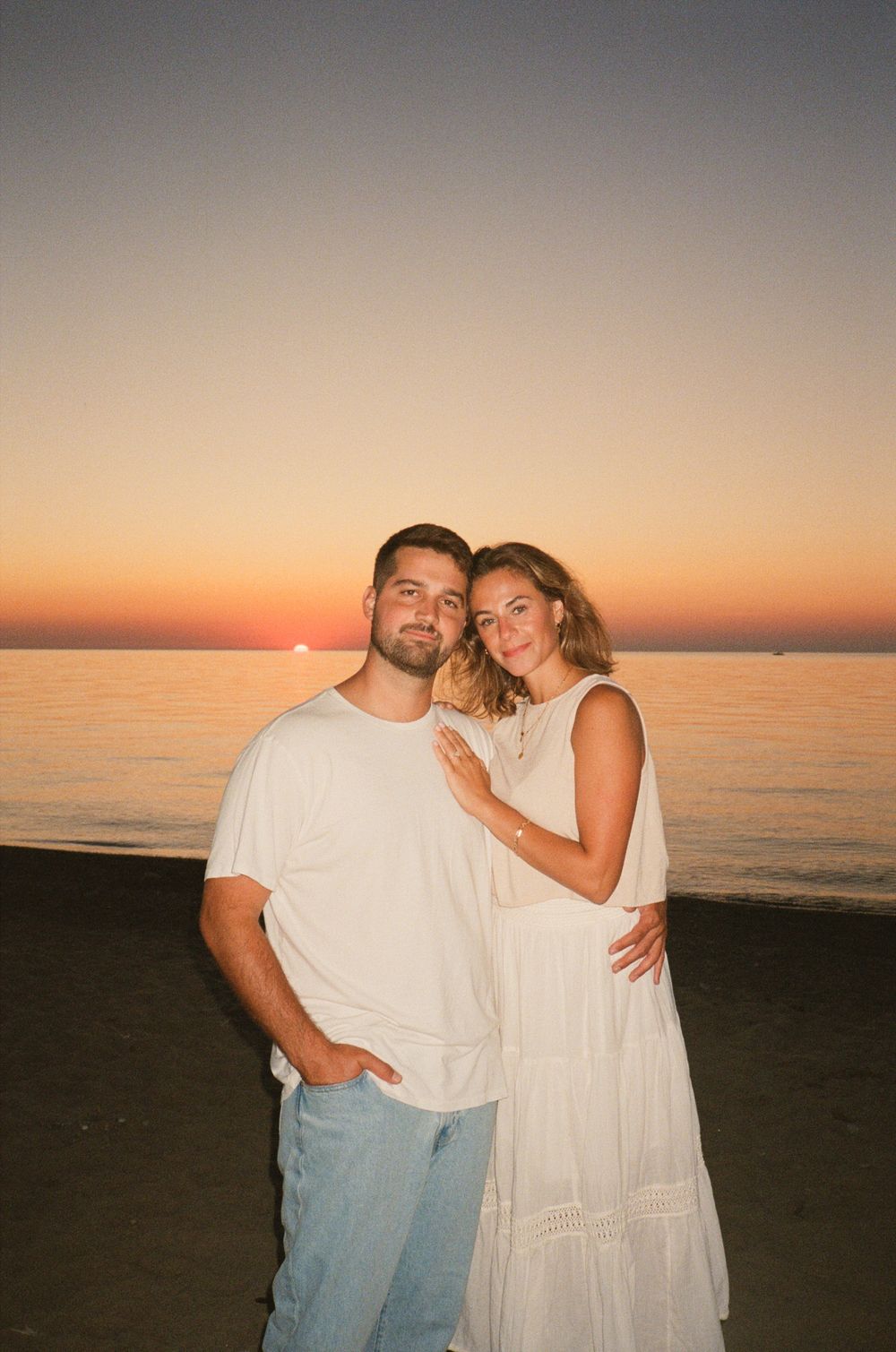 A couple in white clothing embraces on a beach at sunset with the ocean horizon glowing in warm golden light.