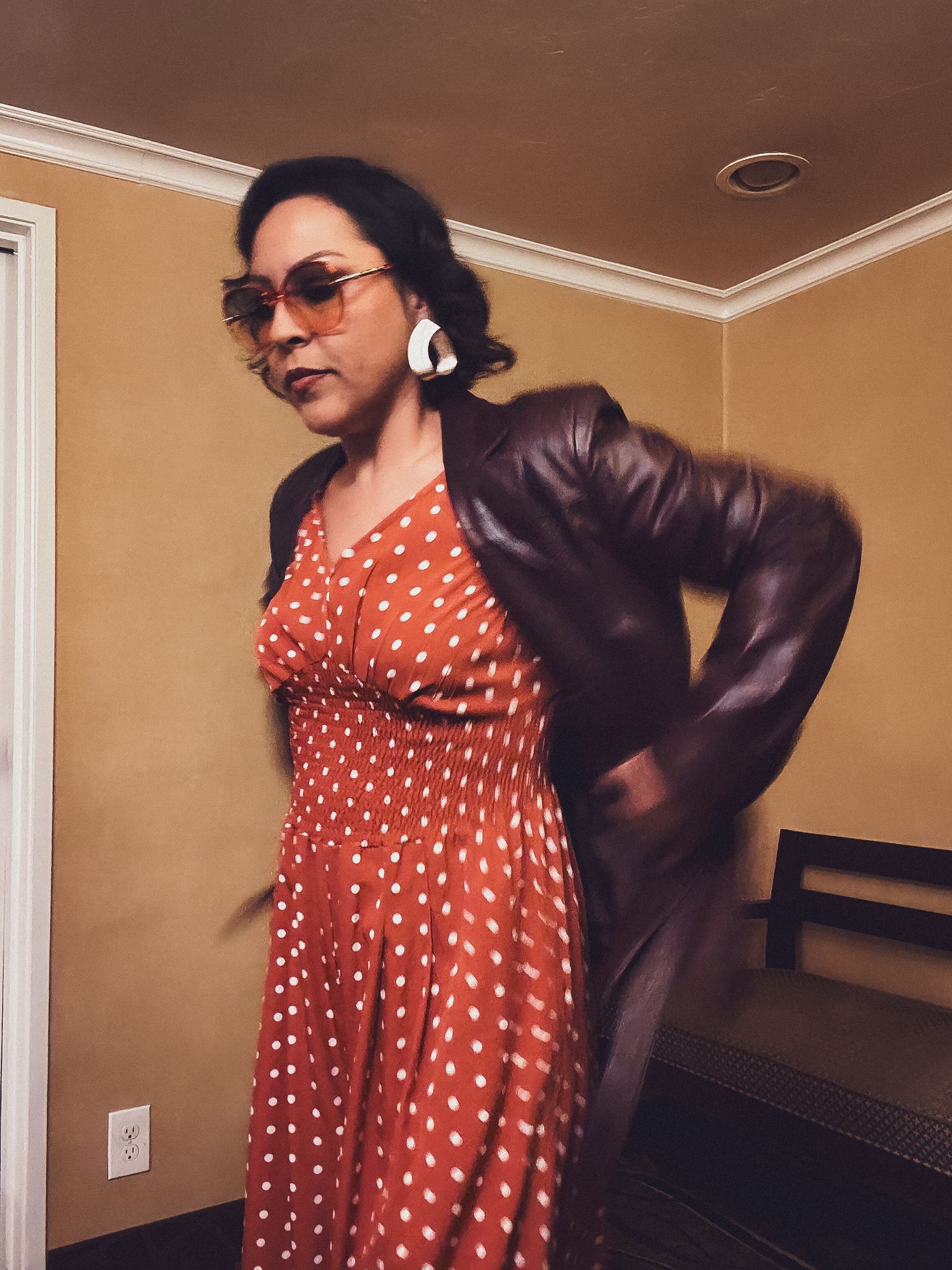 A person wearing a red polka dot dress and black jacket poses playfully in a room with beige walls.