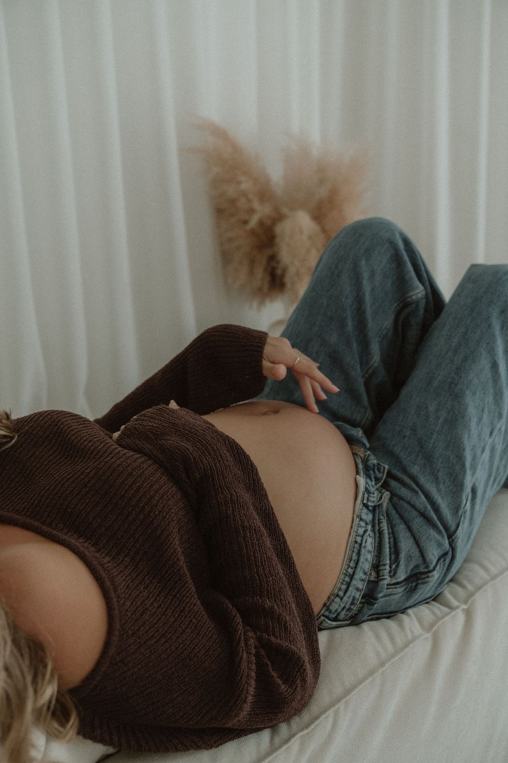 Pregnant woman lying down, wearing brown sweater and jeans, gently touching her baby bump.