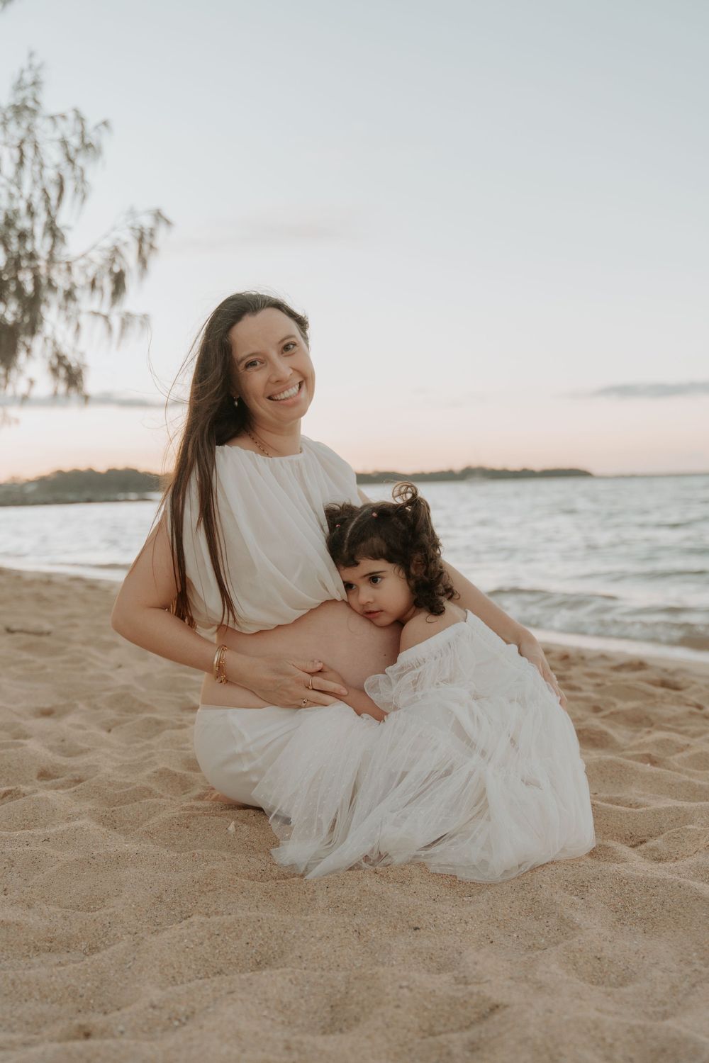 Pregnant woman in white dress smiling on beach with young girl hugging her baby bump at sunset.