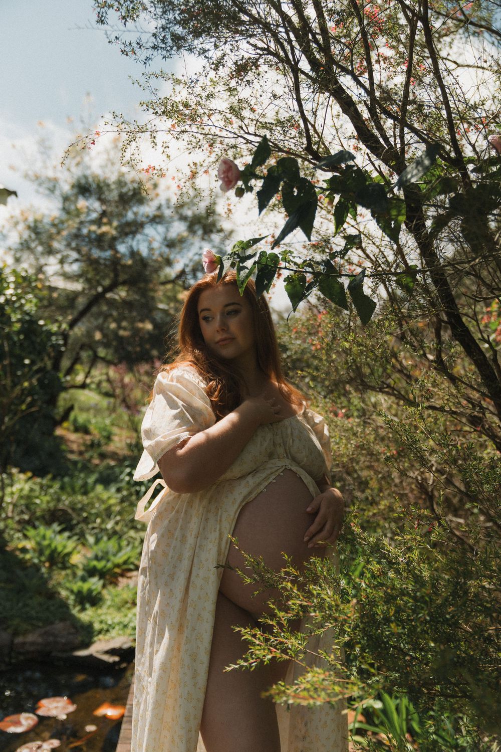 Pregnant woman in flowy white dress holding belly, wearing floral crown, standing in lush green garden with blooming trees.