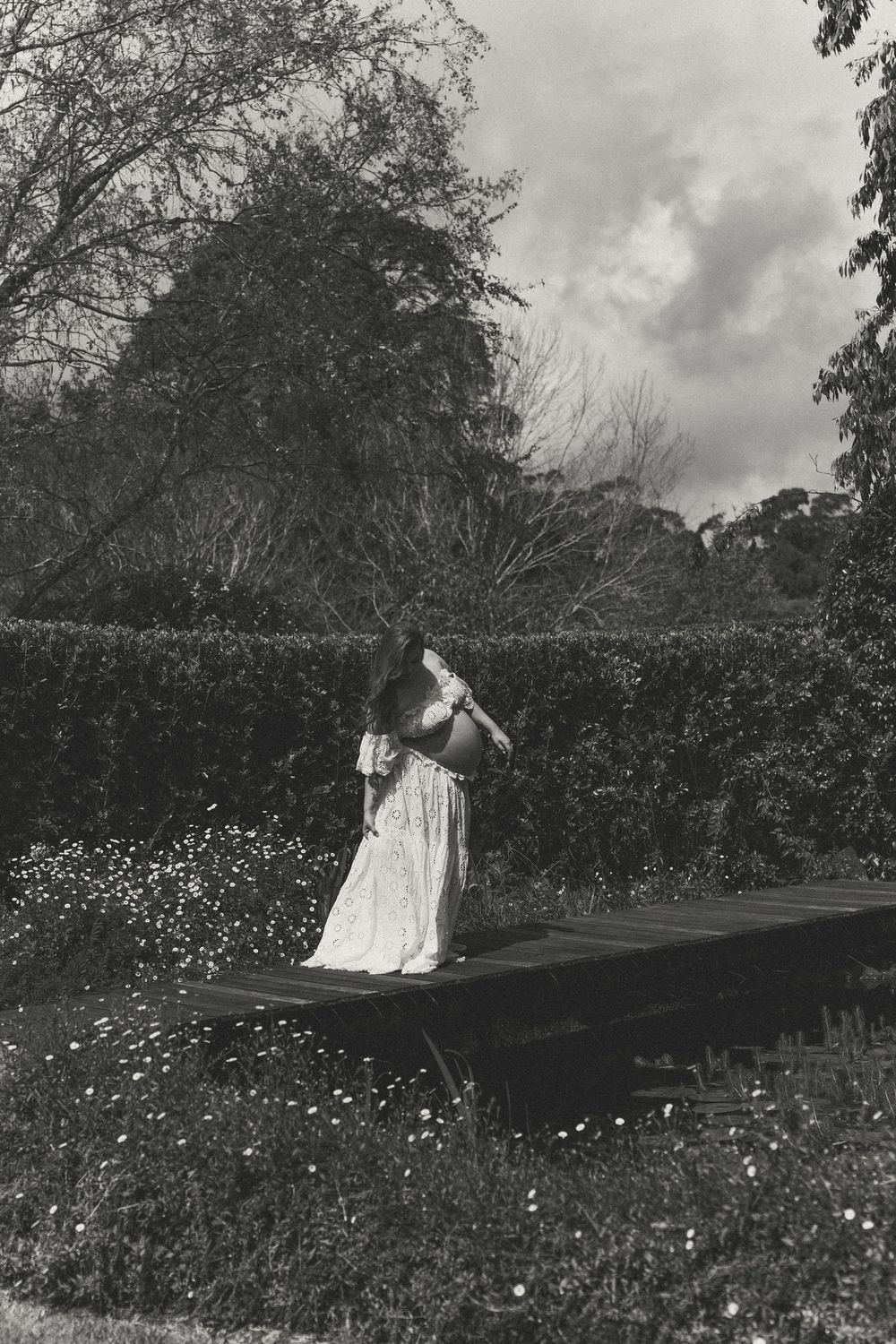 Black and white photo of a woman in a white dress standing on a wooden platform surrounded by lush garden hedges.