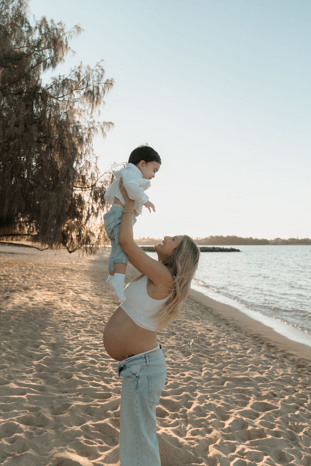 Pregnant woman in white crop top and jeans lifting baby on sandy beach at sunset with trees and calm water behind.