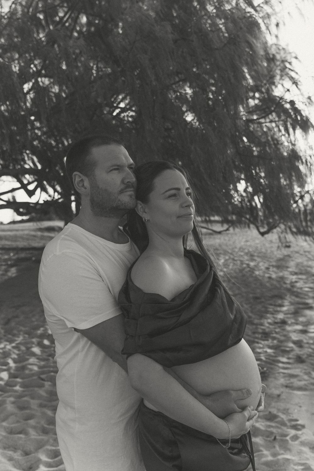 Black and white photo of a couple embracing on a beach, woman showing pregnant belly.