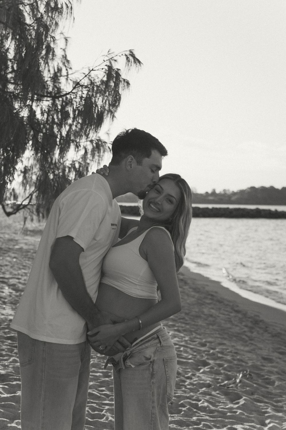 Black and white photo of couple embracing on beach, man kissing pregnant womans head.