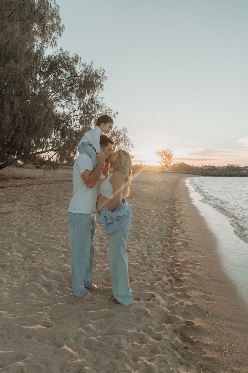 Family of three enjoying a golden sunset on a sandy beach, with child on father's shoulders and mother leaning in close.