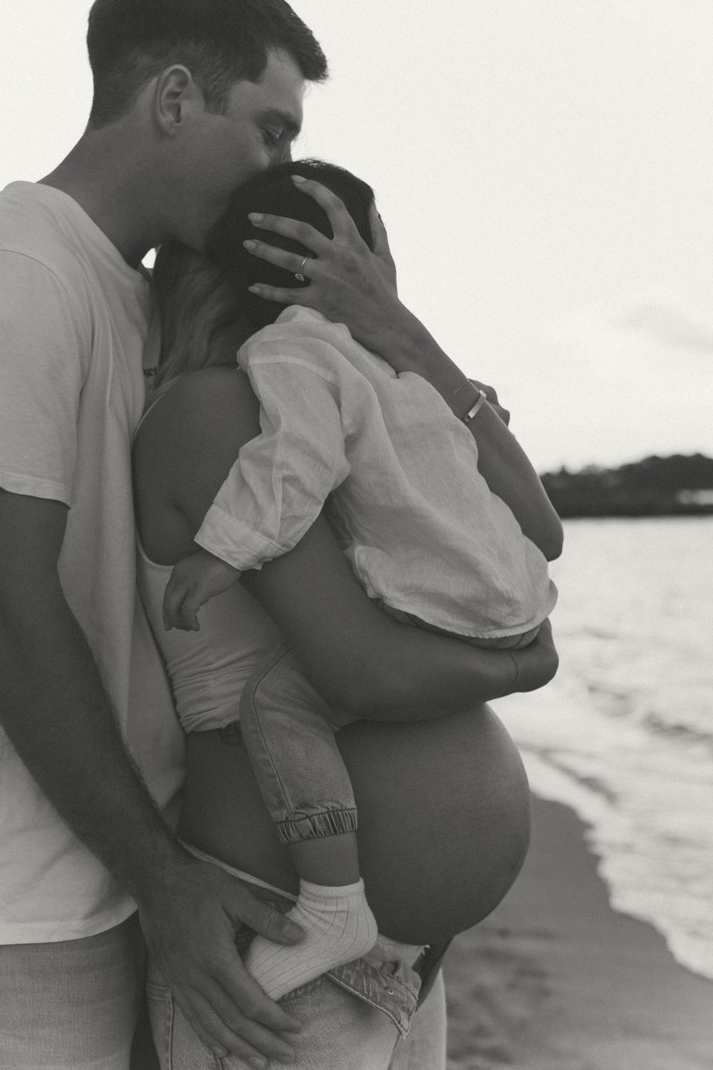 Black and white beach maternity photo of couple embracing, man kissing woman's head while she holds baby bump near ocean.