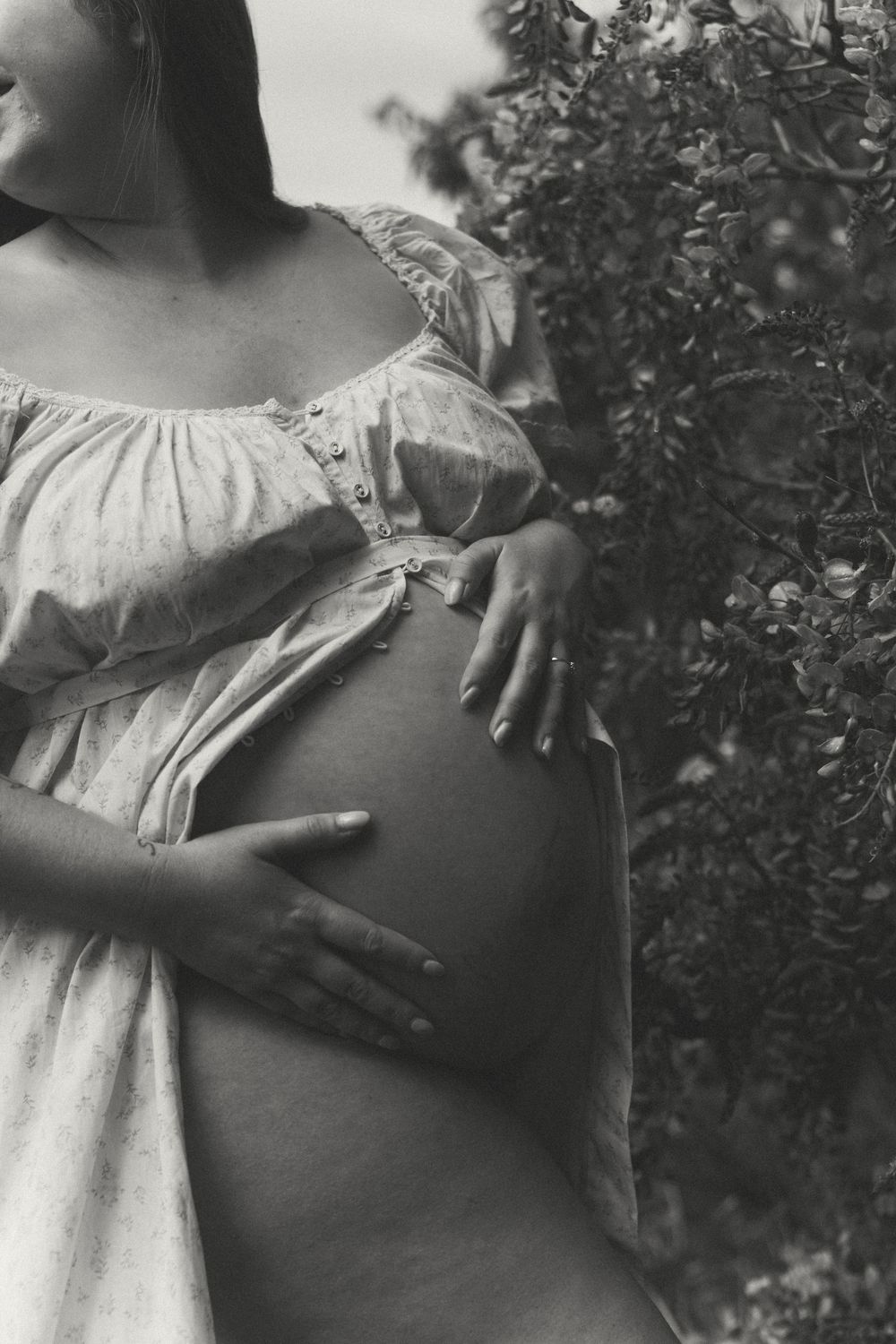 Black and white maternity photo of pregnant woman in flowy dress cradling baby bump outdoors near lush greenery.