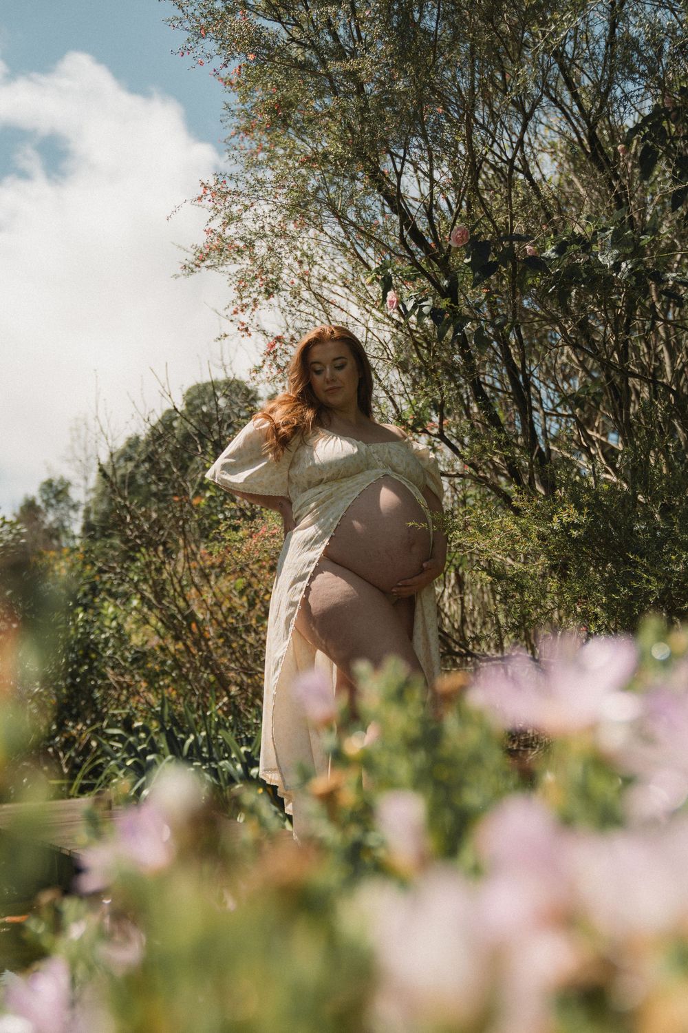 Pregnant woman in white dress standing among wildflowers and trees on a sunny day.