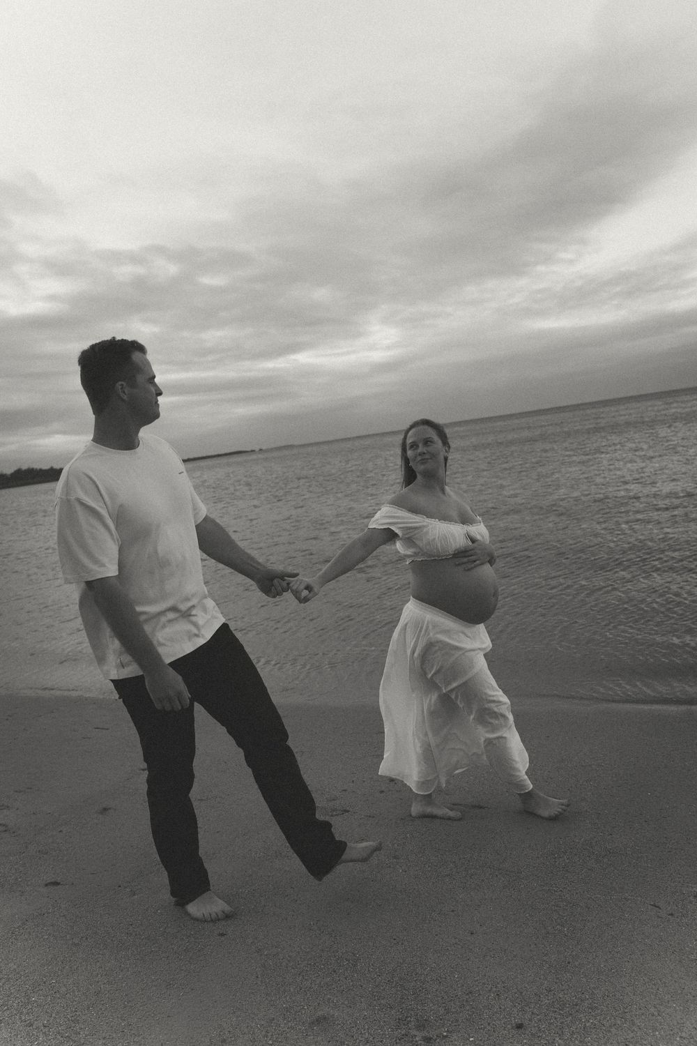 Black and white photo of pregnant couple holding hands and walking along a scenic ocean beach.
