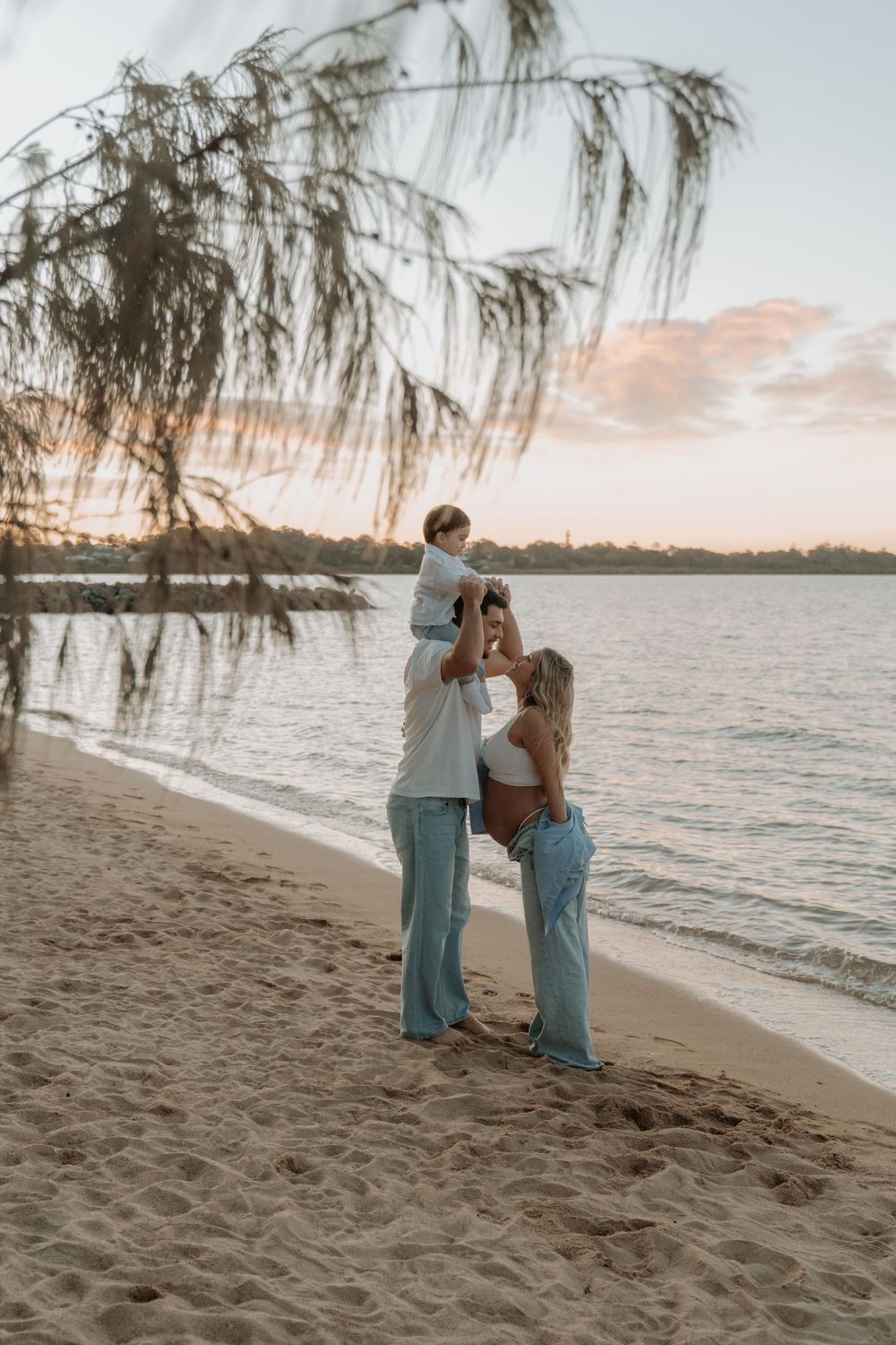 Family of three enjoying sunset on sandy beach, father lifting child near water.