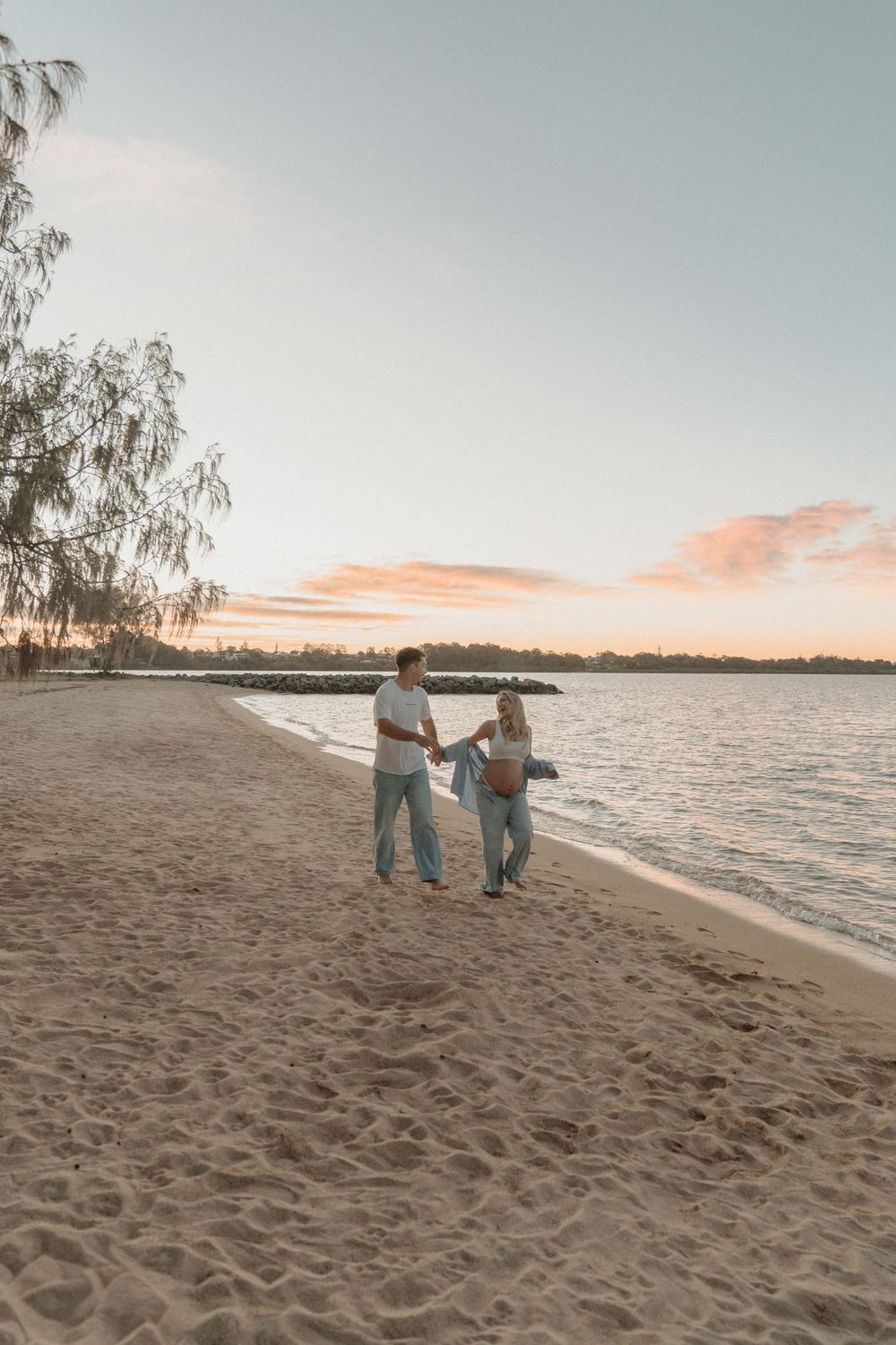 Expecting couple walking hand in hand along sandy beach during beautiful pastel sunset.