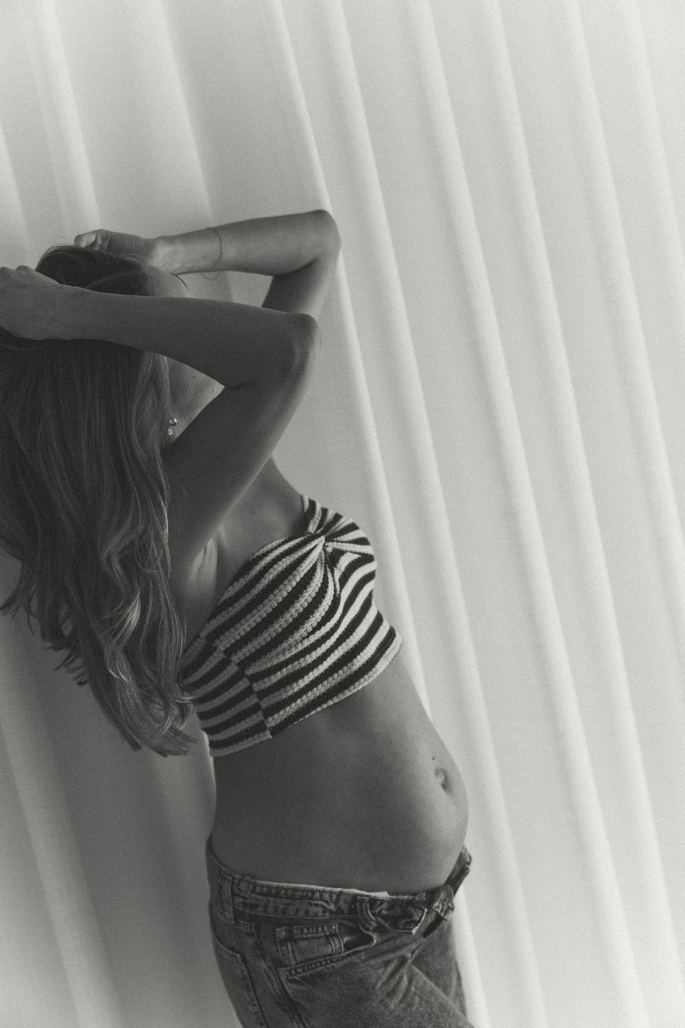 Black and white photo of a woman in striped crop top and jeans posing against white vertical blinds.