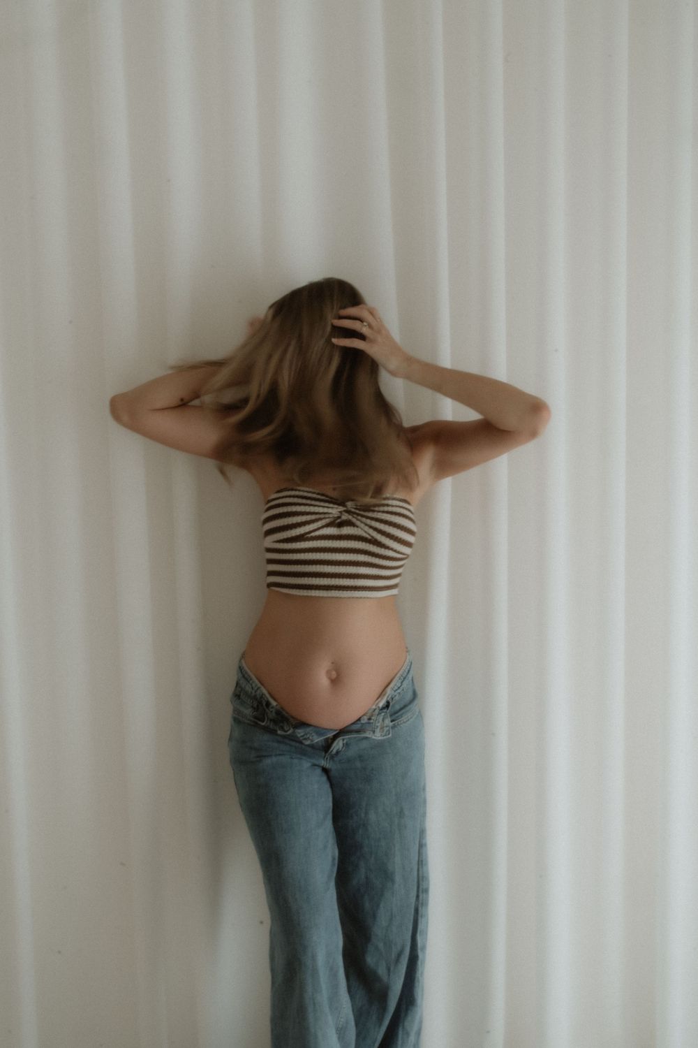 Woman in striped crop top and low-rise jeans standing against white curtain backdrop with hands in hair.