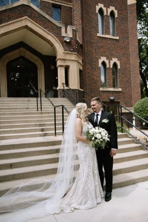 Newlyweds pose on church steps with bride in lace gown holding white bouquet.