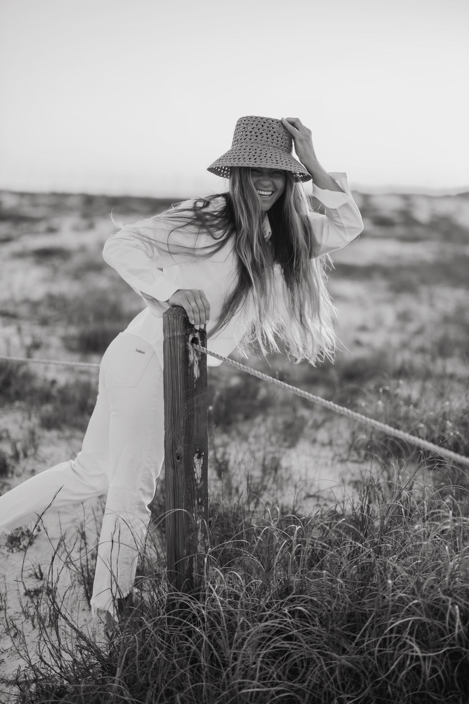 Ethereal black and white portrait of someone in a flowing dress and hat standing in tall grass.