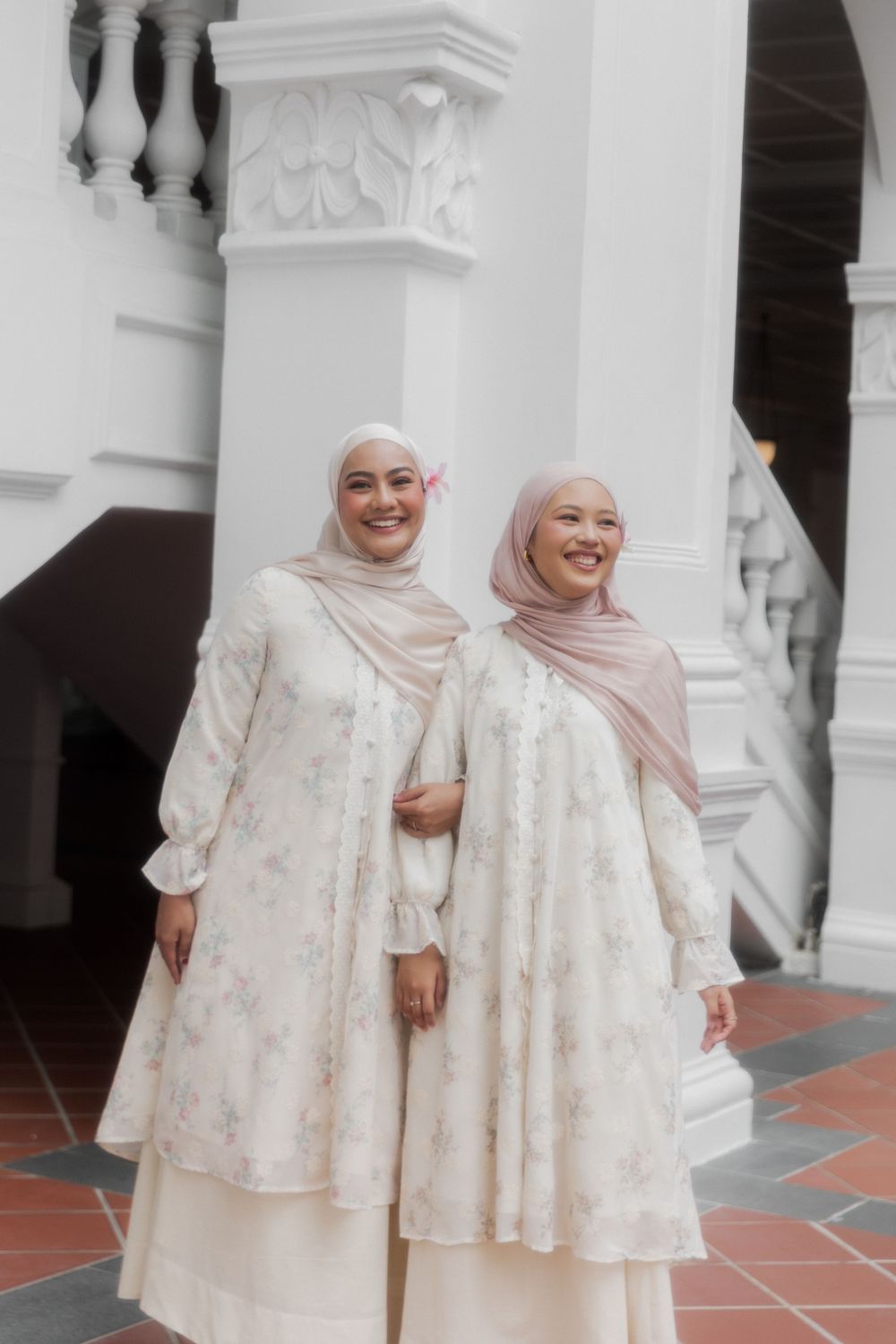 Two women in matching white floral dresses and hijabs pose together in front of classical white columns.
