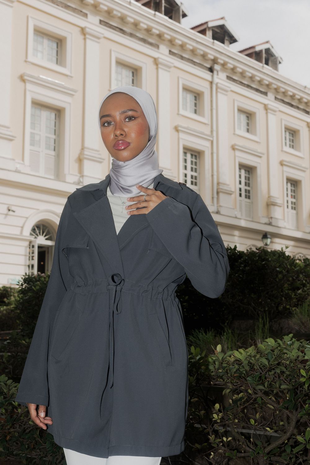 Woman in grey jacket and white hijab poses elegantly against classic architectural backdrop.
