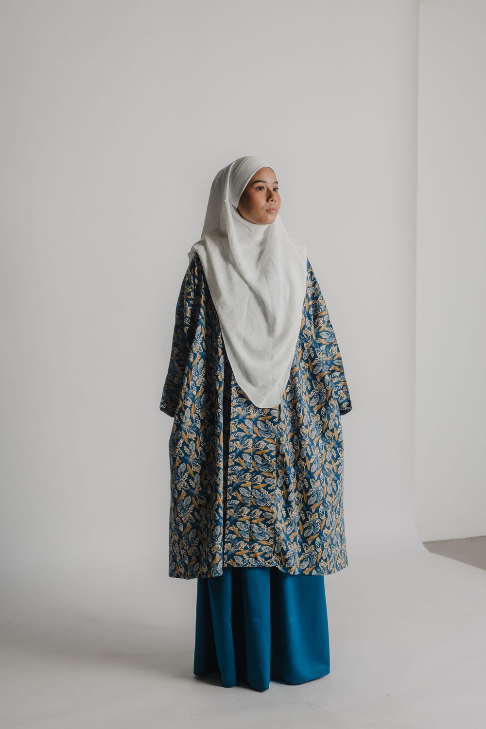 Person wearing patterned blue outfit and white hijab poses against minimal white background.