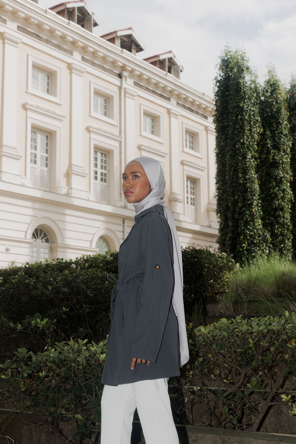 Person in gray blazer and white hijab poses in front of classic white colonial building with greenery.