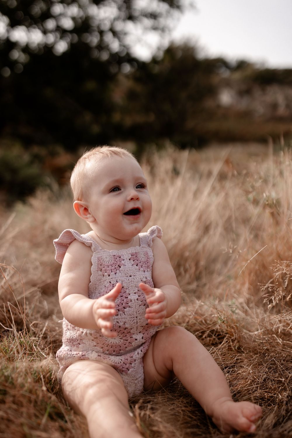Baby in floral romper sitting in tall grass during golden hour photography session.