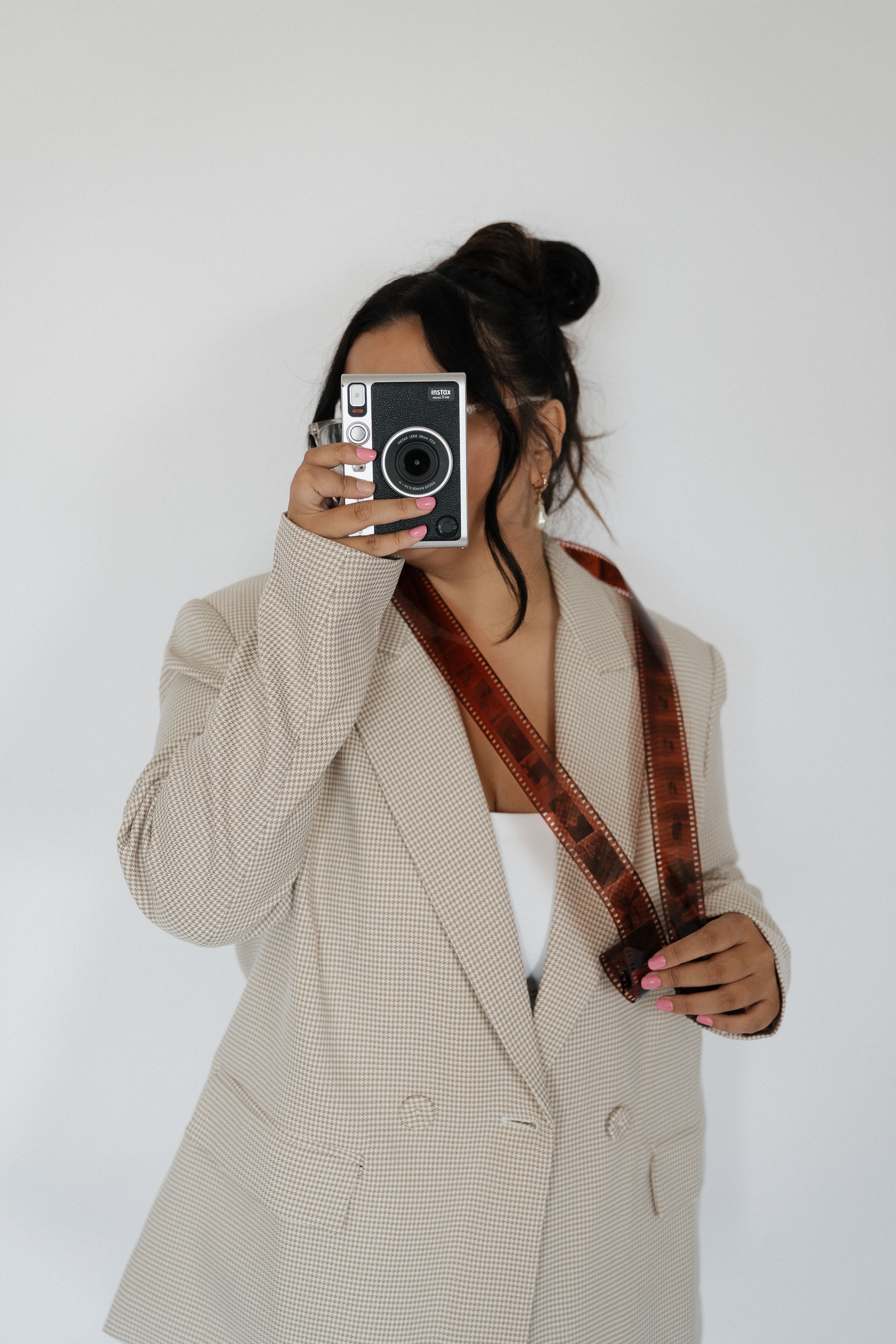 A photographer wearing a beige blazer and glasses smiles while holding a professional camera in a studio.