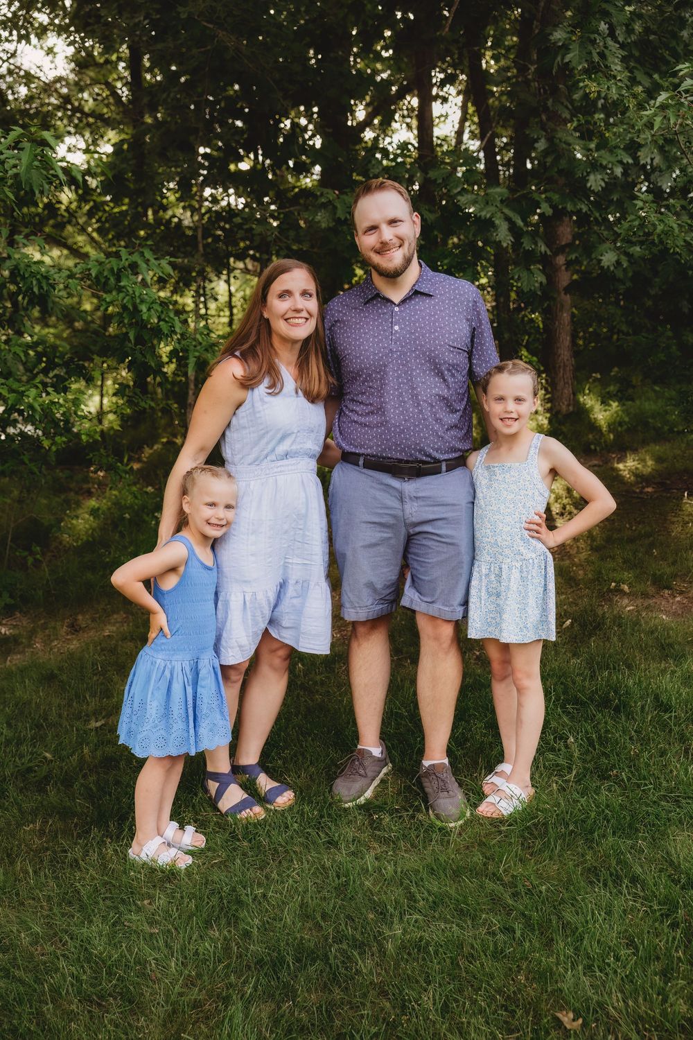 A happy family of four poses outdoors on green grass with trees in the background on a sunny day.