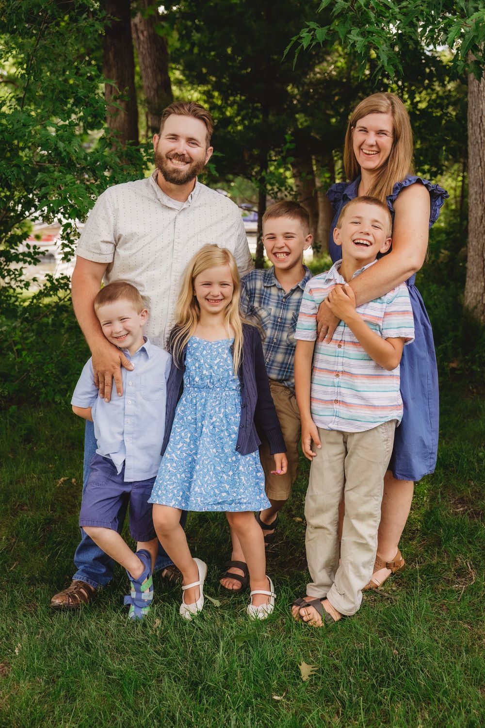 Happy family of six posing outdoors on green grass surrounded by lush trees, parents with four smiling children.