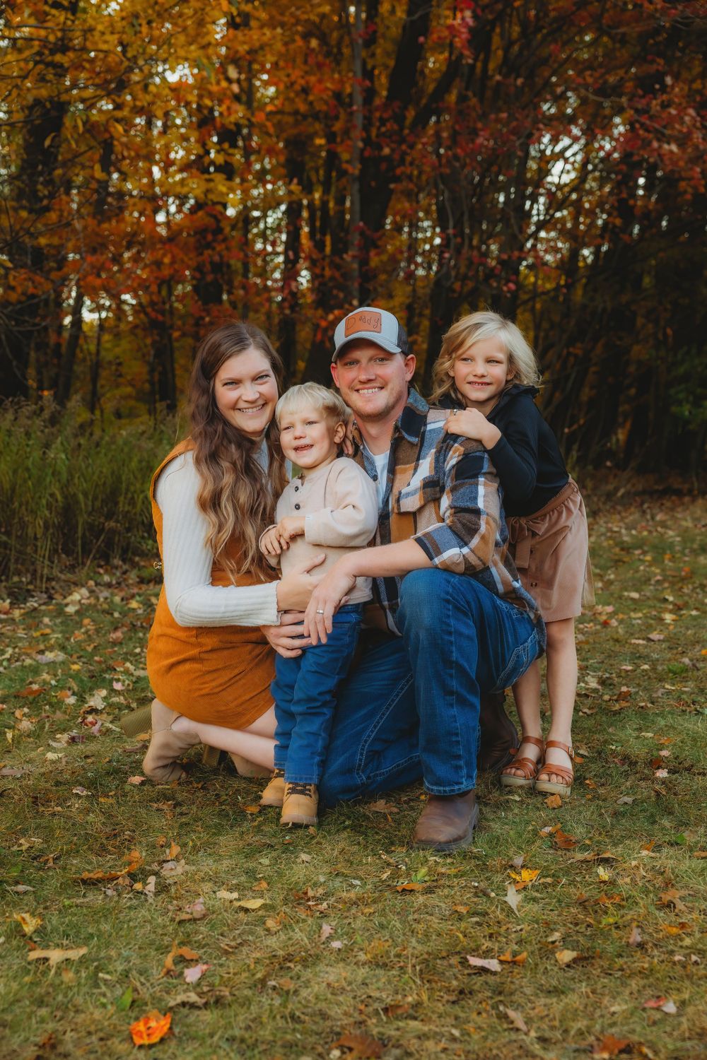 Family sitting together on grass during autumn photoshoot with colorful fall foliage in background.