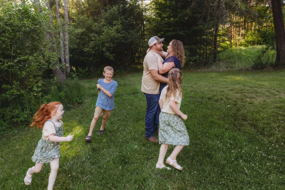 Family of five playing outdoors on green grass surrounded by trees during golden hour.
