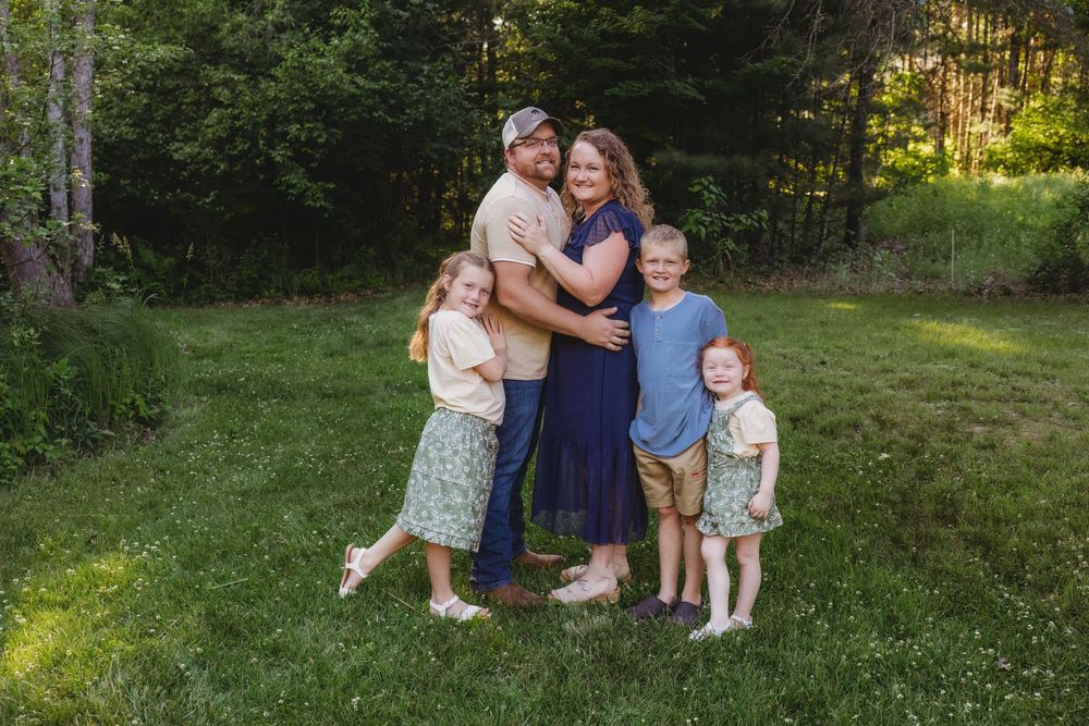 A happy family of five poses together on a green lawn surrounded by lush trees during a warm outdoor photo session.
