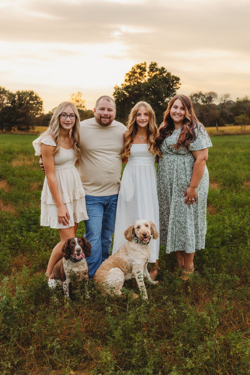 A family with two dogs poses together in a field during golden hour wearing coordinated white and floral outfits.