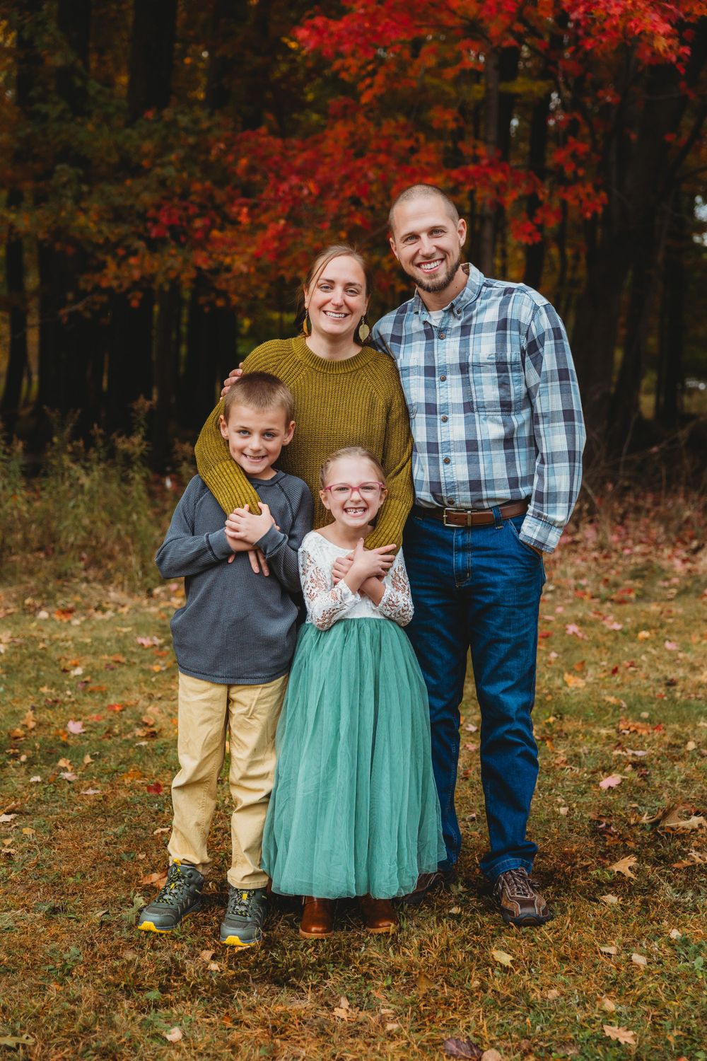 A family portrait featuring bright red autumn trees in the background and coordinated casual outfits.