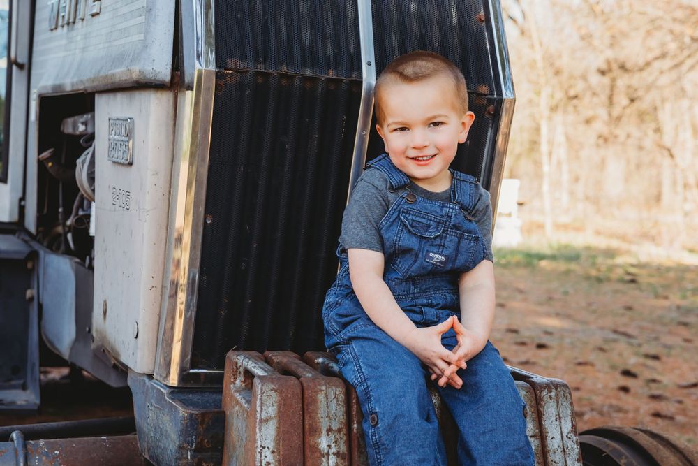 A small child sits smiling in denim overalls on vintage farm equipment.