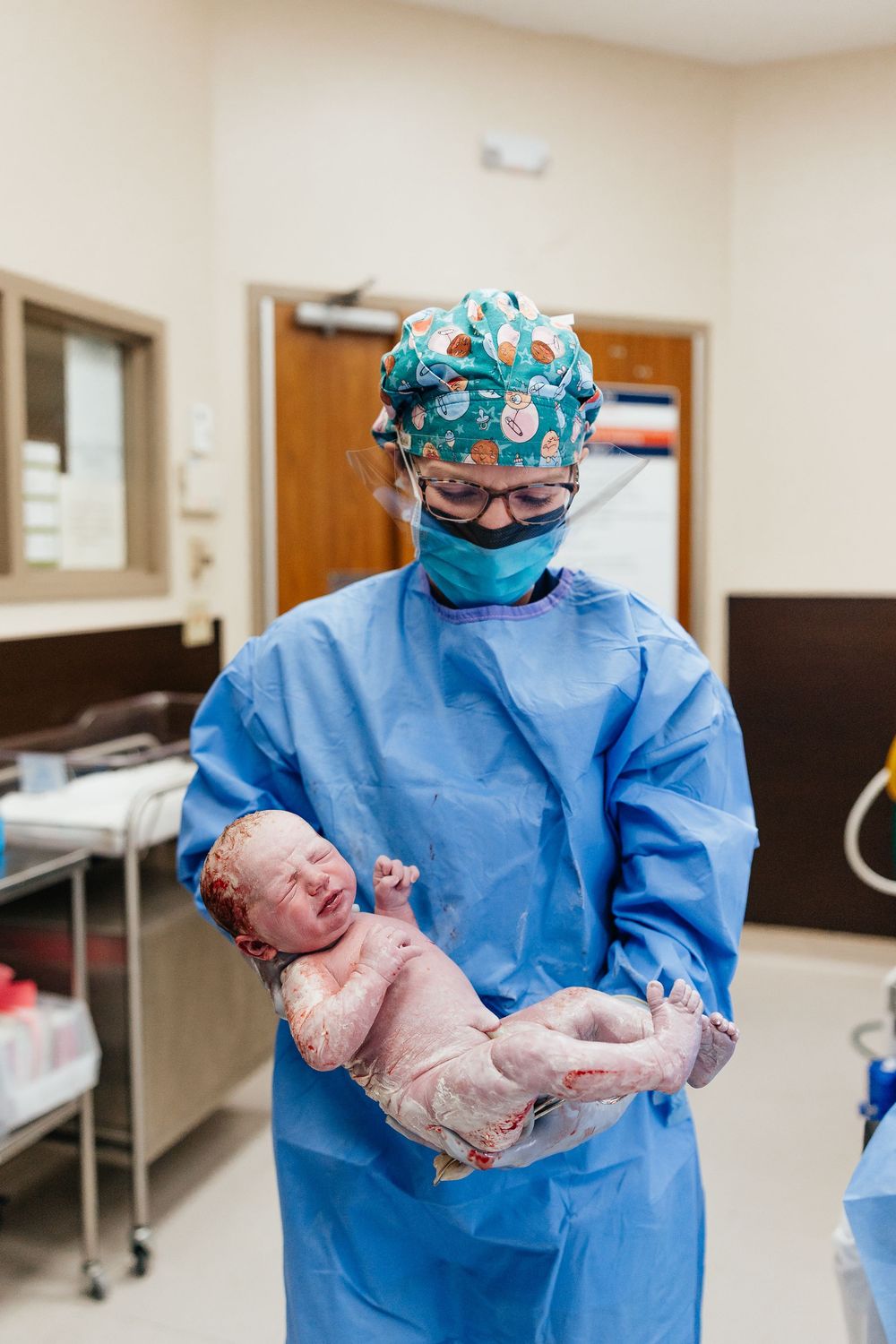 Medical professional in blue scrubs and protective gear holds newborn baby in hospital delivery room.