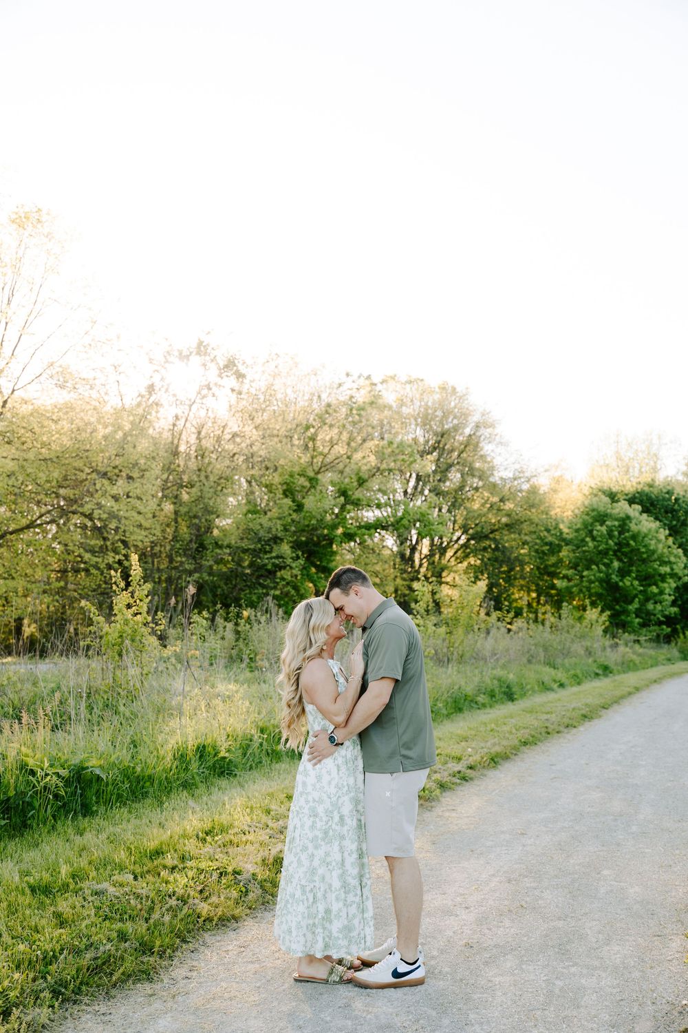 Romantic couple embraces on a dirt path surrounded by lush greenery during golden hour sunset.