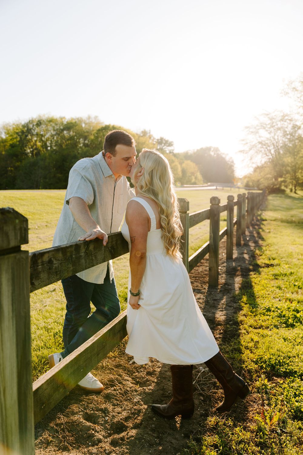 A romantic couple shares a tender moment along a rustic wooden fence at sunset in a pastoral countryside setting.