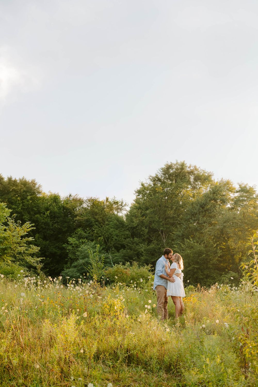 A romantic couple stands together in a sunlit meadow filled with wildflowers and tall grass at golden hour.