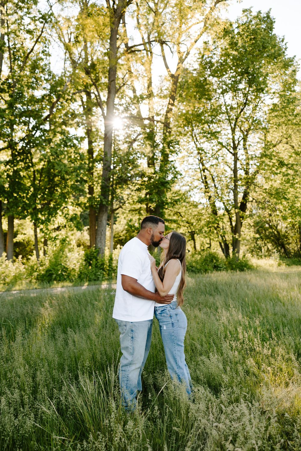 A couple shares a romantic moment in a sunlit meadow with tall grass and trees during golden hour.