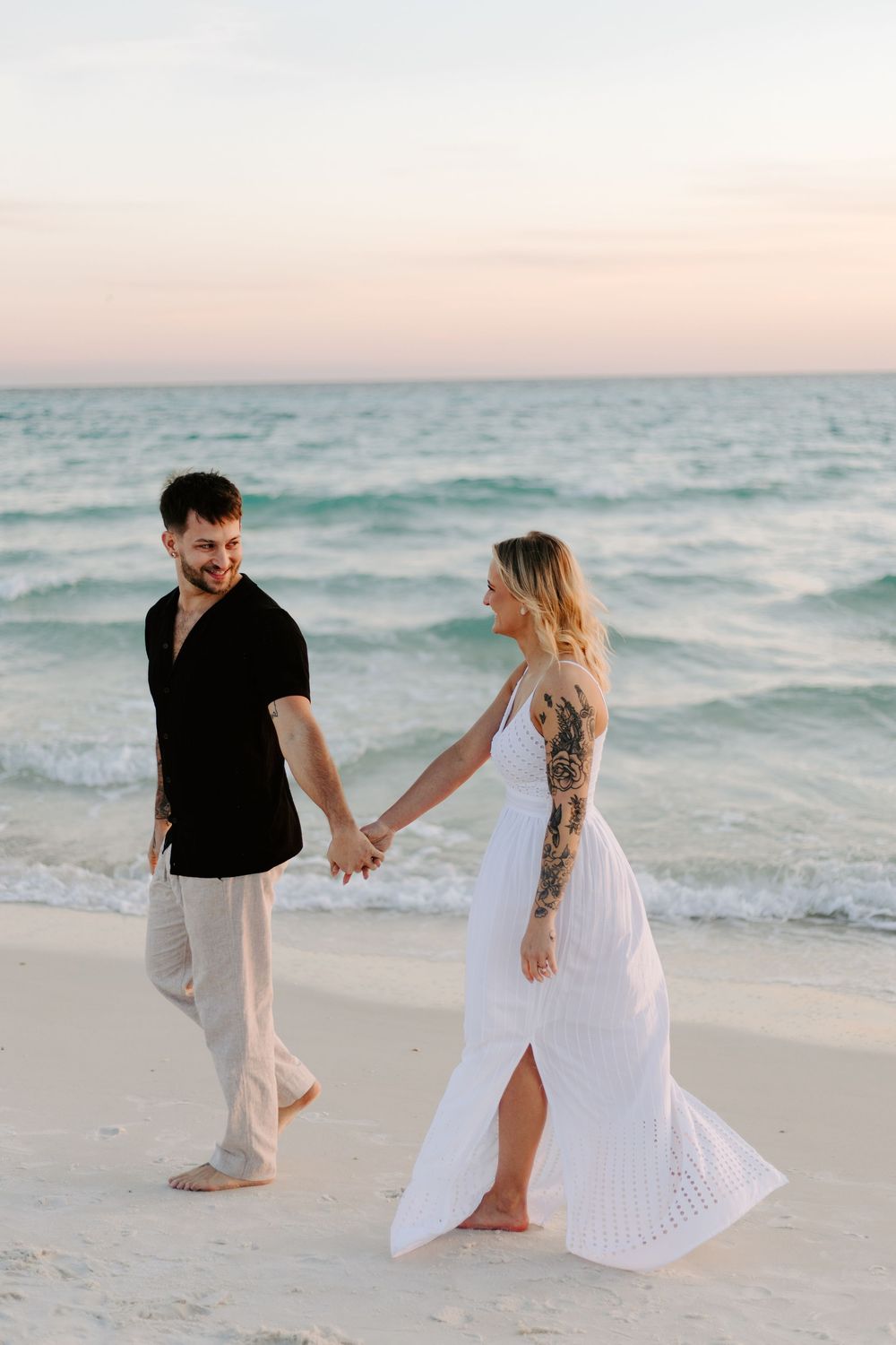 Couple walking hand in hand along a serene beach at sunset with waves rolling in and white sand beneath their feet.