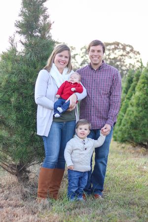 Family of four posing outdoors at a Christmas tree farm, parents holding young children, wearing festive holiday attire.