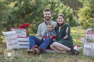Family of three poses outdoors at Christmas tree farm with festive holiday signs and decorations for seasonal portrait.
