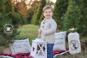 Smiling toddler boy in cream sweater poses at Christmas tree farm with holiday lanterns and festive burlap sacks.